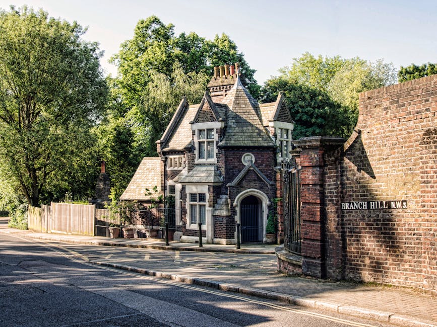 A narrow street at the corner of a residential area in Blackheath, showing an ornate, old-style brick house with pointed turrets, decorative stonework, and multiple windows, some with white frames. The house is partially obscured by green leafy trees, with a wooden fence and a metal gate visible on the left side. The pavement runs along the front of the property, with a curb separating it from the asphalt road. A street sign on a brick wall reads 'Branch Hill NW3,' indicating the location. In the foreground, part of a city van or lorry used for home relocation and furniture transport is visible, positioned near the driveway to facilitate moving services by Man and Van Blackheath. The scene is illuminated by natural daylight, casting shadows on the brick wall and street pavement, capturing the typical quiet and historic character of a Blackheath neighbourhood during a clear day.