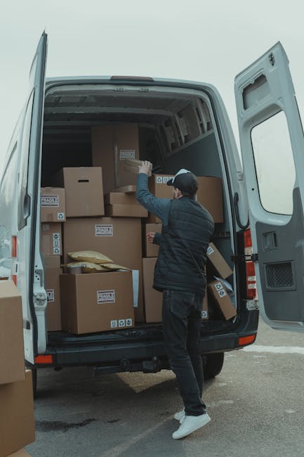 A man with a beard, wearing a white T-shirt and a black cap, is loading a large cardboard box into a moving van parked outside a building. The van's interior is filled with multiple unpacked cardboard