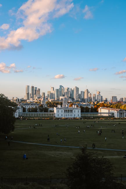 An outdoor scene showing a wide, grassy park area with multiple people walking and gathering on the lawns, some sitting on the grass. In the foreground, there are a few trees and a pathway. Behind the
