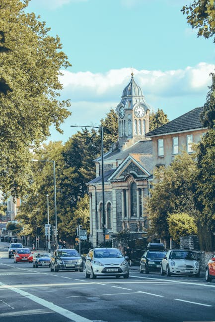 An aerial view of a busy urban street in Blackheath, showing parked cars along the roadside, including a line of delivery vans and smaller vehicles, with pedestrians walking on both the pavement and crossing the road at designated crosswalks. Several trees with lush green foliage line the street, providing shade and a natural contrast to the nearby buildings, which include a mix of modern and historic architecture such as brick and stone facades. Some buildings feature commercial signage, while others appear residential or office spaces. The street is part of a typical city centre environment suitable for house removals and furniture transport, with the scene capturing the ongoing loading and unloading process in an area serviced by Man and Van Blackheath, specialists in removals and packing for house relocations. Bright daylight highlights the details of items being moved, such as boxes and furniture, amidst the urban setting.