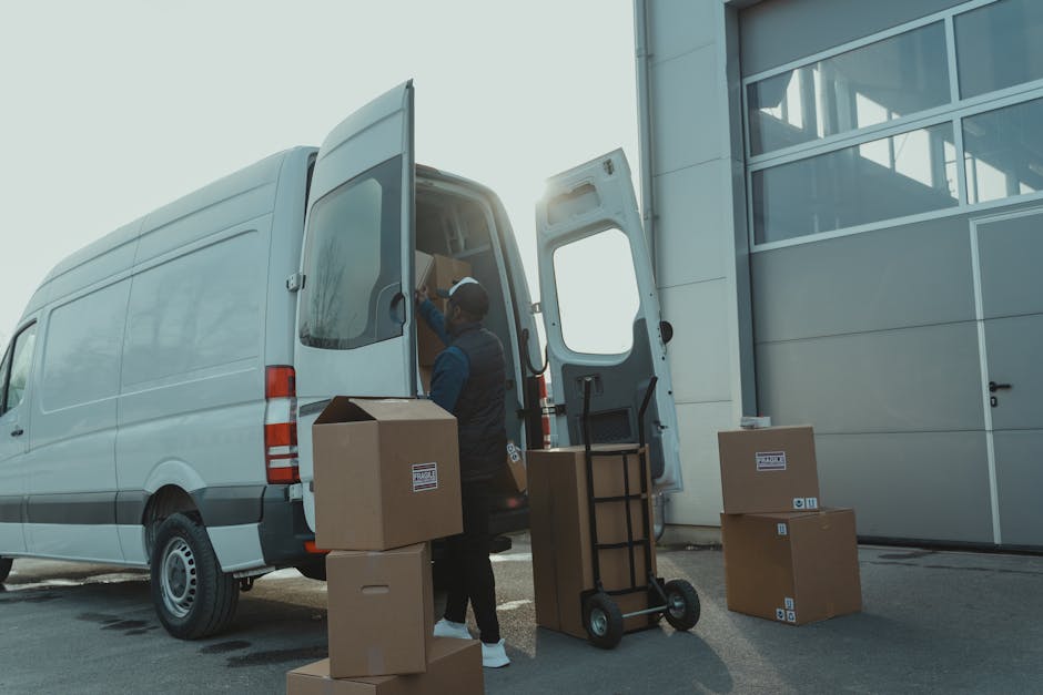A worker from Man and Van Blackheath is seen loading and unloading cardboard boxes from a white delivery van parked outside a modern building with large glass windows. The activity takes place on a paved surface, with several packed boxes placed on the ground near the van and a small hand truck positioned for facilitating the furniture transport process. The worker, dressed in dark clothing and wearing a cap, is inside the van reaching for items or preparing to load or unload. The sunlight casts a glow, highlighting the organized packing materials, which include cardboard boxes sealed with packing tape and some wrapped in plastic for protection during home relocation. The scene captures the typical logistics involved in a professional removals service, emphasizing careful handling and strategic positioning of boxes for efficient furniture transport and packing and moving operations, as provided by Man and Van Blackheath.
