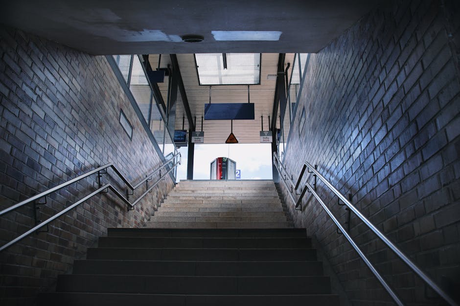 A metal staircase with black steps and silver handrails leading up from an underground station platform to an outdoor area at Blackheath Station. The stairs are flanked by brick walls with some small rectangular signage attached, and an overhead structure with a skylight allows natural light to illuminate the stairwell. At the top of the stairs, there is a clear view of a platform with signage, and a red public telephone booth is visible in the background. The scene suggests an urban setting suitable for facilitating home relocation or furniture transport, with the stairwell possibly used during moving logistics managed by services like Man and Van Blackheath, offering efficient access for loading and unloading items during packing and moving tasks.