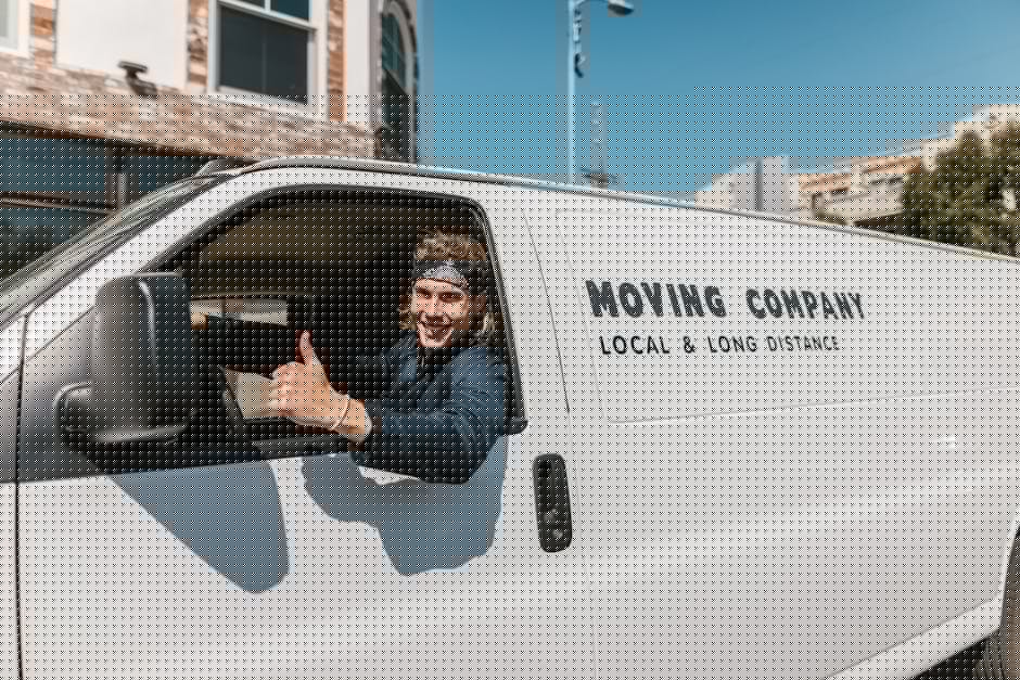 A worker from Man and Van Blackheath is seen loading and unloading cardboard boxes from a white delivery van parked outside a modern building with large glass windows. The activity takes place on a paved surface, with several packed boxes placed on the ground near the van and a small hand truck positioned for facilitating the furniture transport process. The worker, dressed in dark clothing and wearing a cap, is inside the van reaching for items or preparing to load or unload. The sunlight casts a glow, highlighting the organized packing materials, which include cardboard boxes sealed with packing tape and some wrapped in plastic for protection during home relocation. The scene captures the typical logistics involved in a professional removals service, emphasizing careful handling and strategic positioning of boxes for efficient furniture transport and packing and moving operations, as provided by Man and Van Blackheath.
