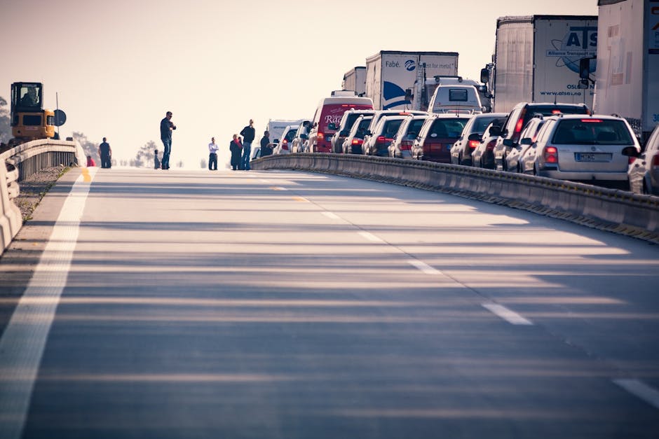 A line of stationary vehicles, including vans and trucks, parked along the side of a busy road, with several people standing and walking on the pavement near the vehicles. The scene appears to be in a