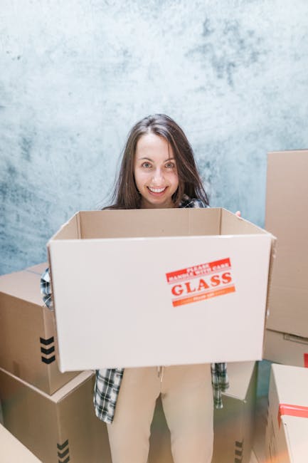 A woman with long brown hair, smiling, holding an empty cardboard moving box labeled 'fragile glass' in a room with a light blue textured wall. She is standing among other packed cardboard boxes, some sealed with tape, which are stacked on the floor and against the wall. The boxes vary in size and are made of brown corrugated cardboard, with some featuring printed or taped labels. The woman is dressed in casual clothing, including a plaid shirt and beige trousers. The scene captures a home relocation or packing process, with the interior environment illuminated by natural or soft lighting. Man and Van Blackheath specializes in furniture transport and packing and moving services, as reflected in the context of this scene.