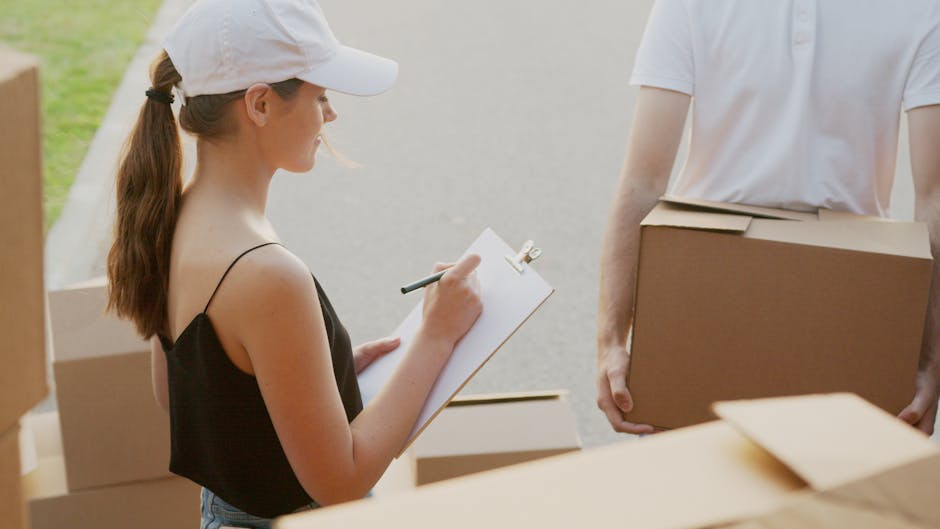 A woman with light skin, wearing a white baseball cap and a black sleeveless top, is holding a clipboard and pen, taking notes during a home relocation process. She is standing outdoors on a paved area near a property entrance, with natural light illuminating the scene. Behind her, several cardboard boxes of various sizes are visible, some opened and others taped closed, indicating packing and moving preparations. To her right, a man with light skin wearing a white T-shirt is holding a medium-sized cardboard box, possibly about to load it onto a vehicle or carry it into the property. The setting suggests a furniture transport or packing and moving operation, with the background showing a residential environment. This scene involves activities typical of professional removals, such as inventory checking, packing, and loading, and reflects elements of home relocation logistics that are often managed by services like Man and Van Blackheath.