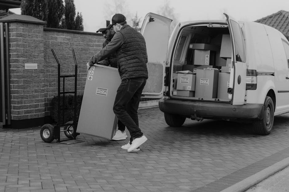 A professional mover from Man and Van Blackheath is seen outside a building during a home relocation, dressed in a dark puffer jacket and white cap, standing on a paved area next to a white van filled