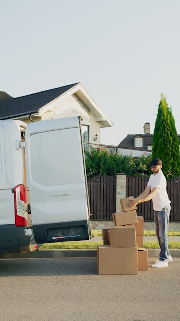 A man wearing a white t-shirt and glasses is seen loading cardboard boxes onto the pavement next to a white van with its rear doors open, prepared for a home relocation. The boxes are of various sizes, with some stacked on the ground and others being carried by the man. The scene is set on a residential street with a house featuring a sloped roof, a chimney, and surrounding greenery, including a tall tree and a wooden fence. Inside the van, visible packing materials such as brown paper and cardboard boxes indicate packing and moving activities. The lighting suggests daytime, and the setting appears organized, with the man carefully placing boxes in preparation for furniture transport or home removals. The branded location associated with [COMPANY_NAME] and the service category of removals suggests this is part of a professional move or relocation process supported by [COMPANY_NAME], emphasizing efficient loading and transport logistics.