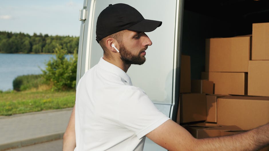 A man wearing a black cap, white t-shirt, and wireless earbuds is standing outside near a white moving van, with the van's open side revealing a stack of uniformly sized cardboard boxes. He is in the process of loading or unloading the boxes, which are arranged on the vehicle's floor inside, with some positioned on top of others. The background includes a paved area and a body of water surrounded by green trees, suggesting an outdoor residential or scenic location typical of house removals or home relocation services. The scene captures the logistics of furniture transport and packing during a removal service, with natural daylight illuminating the area, and the man focused on handling the boxes carefully as part of a professional moving process, such as those offered by Man and Van Blackheath.