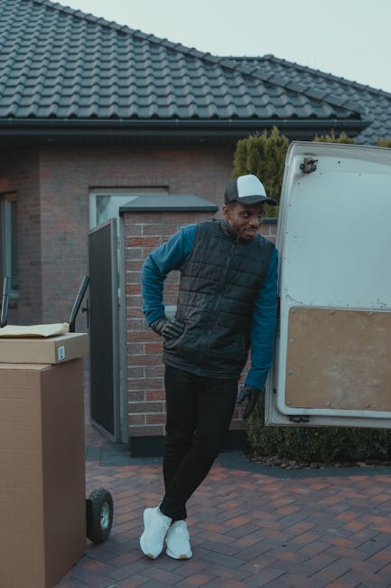 A man wearing a white t-shirt and glasses is seen loading cardboard boxes onto the pavement next to a white van with its rear doors open, prepared for a home relocation. The boxes are of various sizes, with some stacked on the ground and others being carried by the man. The scene is set on a residential street with a house featuring a sloped roof, a chimney, and surrounding greenery, including a tall tree and a wooden fence. Inside the van, visible packing materials such as brown paper and cardboard boxes indicate packing and moving activities. The lighting suggests daytime, and the setting appears organized, with the man carefully placing boxes in preparation for furniture transport or home removals. The branded location associated with [COMPANY_NAME] and the service category of removals suggests this is part of a professional move or relocation process supported by [COMPANY_NAME], emphasizing efficient loading and transport logistics.