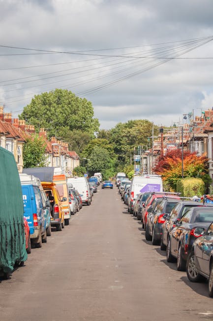 A residential street scene with closely parked cars on both sides, including a variety of makes and models, some covered with protective cloths or plastic wraps, during daylight with a partly cloudy sky. The street is lined with terraced houses featuring brick facades, small front gardens, and pitched roofs. Overhead, there are visible electrical wires stretching across the street. In the foreground, a man from Man and Van Blackheath  is preparing to load furniture or boxes into a van parked on the side of the road, as part of a home relocation or furniture transport process. The environment suggests a typical urban area suitable for house removals and moving services, with ample space for loading and parking in connection with the parking tips for Blackheath Common, BLACKHEATH, as highlighted in the webpage titled 'Removals tips for parking at Blackheath Common, BLACKHEATH.'