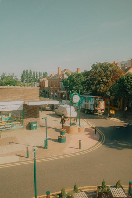 An outdoor scene showing a street in Blackheath during daylight with a clear sky. In the foreground, there is a small paved area with painted yellow road markings and several green metal bollards surrounding potted plants. A woman dressed in casual clothing is walking across the pavement, carrying a small item. To the left, there is a shop with a large glass window displaying assorted goods and a white awning. A green lamppost and an information sign are positioned near the pavements. On the street, a white van and a truck with a partially visible delivery logo are parked along the curb, indicating possible loading activities related to home relocation or furniture transport services. In the background, there are residential buildings with brick exteriors, chimneys, and tree-lined streets, suggesting a suburban neighbourhood. This scene possibly captures a moment during a same-day removals service provided by Man and Van Blackheath, highlighting moving logistics involving loading or unloading of household items in a residential area.