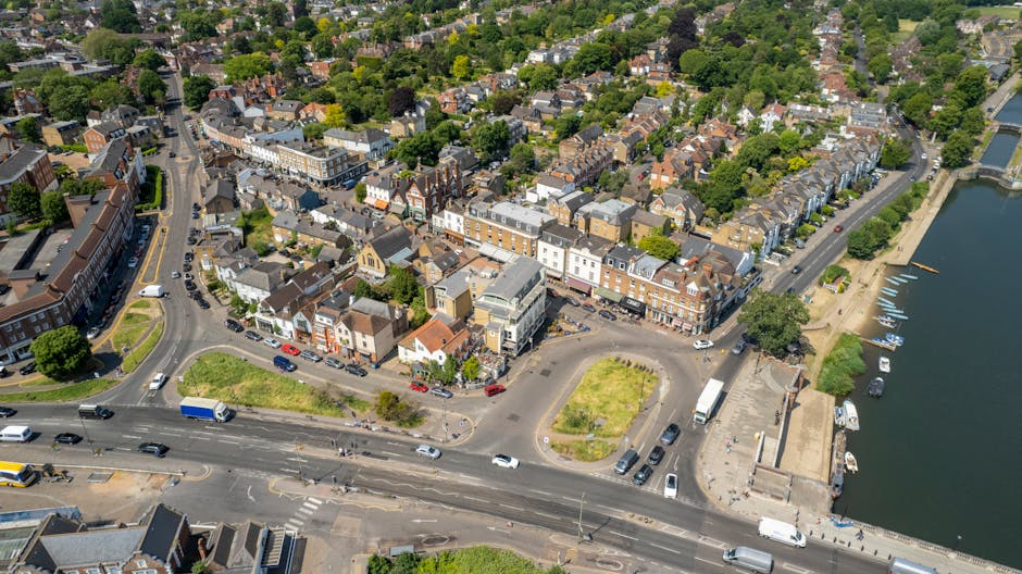 Aerial view of a town featuring a mix of residential and commercial buildings surrounded by green trees and a river. In the foreground, a busy road with cars, trucks, and buses is visible, along with a small roundabout and parking areas. To the right, a paved area with a few parked boats and footpaths runs along the riverbank. Near the center, a cluster of buildings includes shops, offices, and possibly a community centre, with some vehicles parked outside. The densely packed houses with tiled roofs extend into the background, interspersed with patches of greenery and trees in the residential areas. The overall scene is well-lit, showcasing typical UK townscape elements, suitable for discussing house removals, furniture transport, or moving logistics as provided by Man and Van Blackheath, aligned with the content on the page for same day removals in Blackheath.