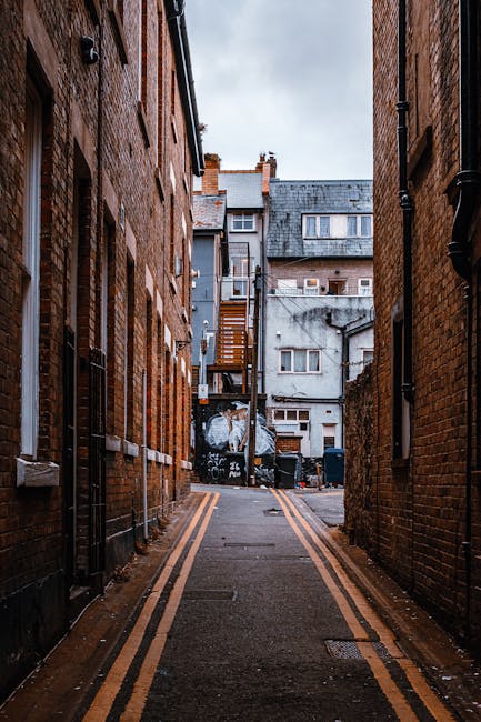 A narrow alleyway between two brick buildings, with a paved surface marked by double yellow lines along each side. The alley is used for home relocation and furniture transport, with several cardboard boxes and moving supplies visible near the entrance. At the end of the alley, there is a small open area with a multi-storey building that has a wooden staircase attached to the exterior, leading to upper floors. The area appears to be part of a communal space or back yard, with some trash bins and graffiti on a wall. The lighting is overcast, indicating daytime conditions typical of urban house removals, and Man and Van Blackheath could be involved in managing such moving logistics in tight street environments. The scene captures the typical challenges of loading and unloading during packing and moving processes in confined urban streets, highlighting the necessity of careful planning and appropriate equipment such as trolleys or straps to facilitate the furniture transport.