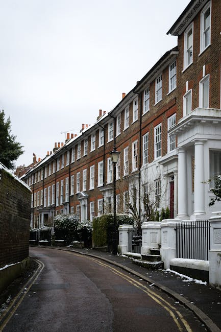 The image shows a narrow residential street in Blackheath during daytime, with a row of traditional brick terraced houses featuring white window frames and decorative columns at the entrance. The street is lined with parked vehicles, including a black van positioned next to the curb, which is being used for home relocation purposes by Man and Van Blackheath. Several cardboard boxes, wrapped in plastic and packing materials, are stacked on the pavement near the van, indicating the packing phase of the furniture transport process. The loading process appears to be ongoing, with a focus on safely loading boxes and furniture into the van, which is parked close to the entrance of one of the houses. The scene is well-lit with natural daylight, and there are sparse patches of snow on the pavement, suggesting cold weather conditions. The setup illustrates a typical house removal operation in a busy urban environment with narrow access, aligning with the advice provided for relocating in SE3 areas with limited street space.