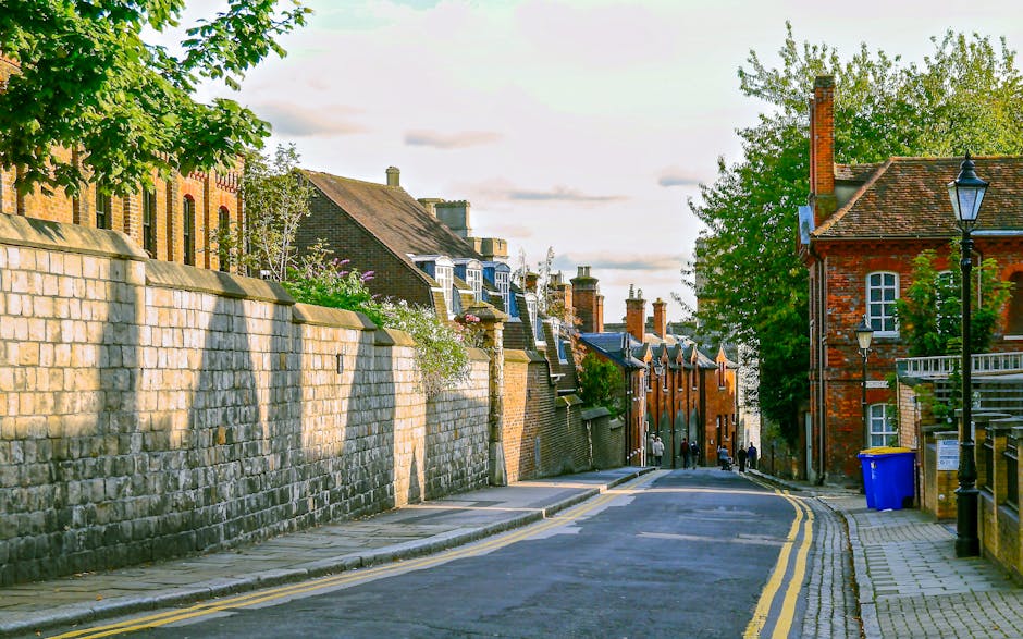 A narrow alleyway between two brick buildings, with a paved surface marked by double yellow lines along each side. The alley is used for home relocation and furniture transport, with several cardboard boxes and moving supplies visible near the entrance. At the end of the alley, there is a small open area with a multi-storey building that has a wooden staircase attached to the exterior, leading to upper floors. The area appears to be part of a communal space or back yard, with some trash bins and graffiti on a wall. The lighting is overcast, indicating daytime conditions typical of urban house removals, and Man and Van Blackheath could be involved in managing such moving logistics in tight street environments. The scene captures the typical challenges of loading and unloading during packing and moving processes in confined urban streets, highlighting the necessity of careful planning and appropriate equipment such as trolleys or straps to facilitate the furniture transport.