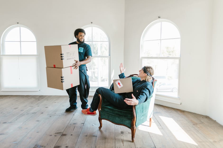 A professional removal service in progress inside a bright, spacious room with natural light coming through four large arched windows. A man with a beard and dark hair, dressed in a blue uniform, is standing on the wooden floor, carrying two cardboard boxes stacked one on top of the other, with red and black packing tape securing the boxes. Opposite him, a woman with glasses and blonde hair, wearing dark clothing, is sitting on an upholstered green armchair, holding and supporting a cardboard box with a white label, and raising her hand to signal for a pause or assistance. The room is empty aside from the furniture and the moving boxes, with smooth white walls and a light-colored wooden floor, illustrating a typical home relocation process involving packing, lifting, and preparing furniture and boxes for transport. Man and Van Blackheath is providing a furniture transport and packing service to facilitate an efficient move.