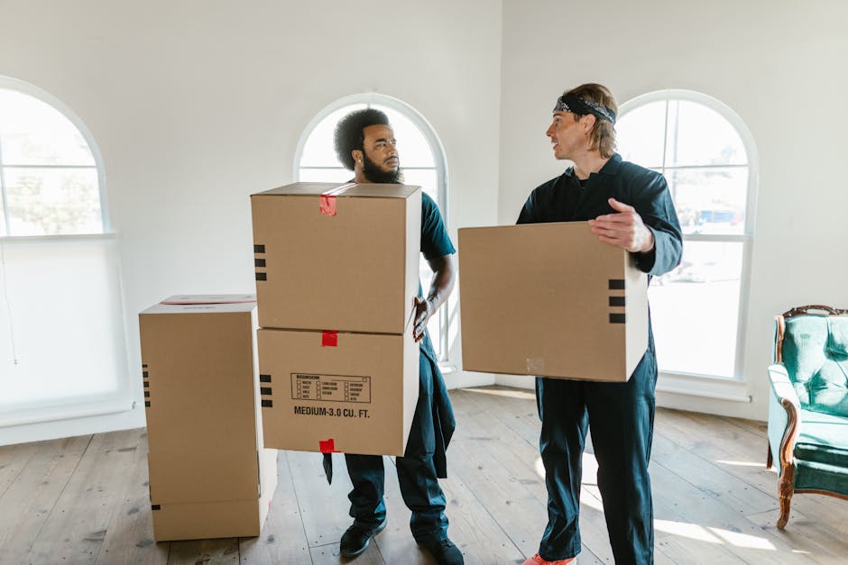 A professional removal service in progress inside a bright, spacious room with natural light coming through four large arched windows. A man with a beard and dark hair, dressed in a blue uniform, is standing on the wooden floor, carrying two cardboard boxes stacked one on top of the other, with red and black packing tape securing the boxes. Opposite him, a woman with glasses and blonde hair, wearing dark clothing, is sitting on an upholstered green armchair, holding and supporting a cardboard box with a white label, and raising her hand to signal for a pause or assistance. The room is empty aside from the furniture and the moving boxes, with smooth white walls and a light-colored wooden floor, illustrating a typical home relocation process involving packing, lifting, and preparing furniture and boxes for transport. Man and Van Blackheath is providing a furniture transport and packing service to facilitate an efficient move.